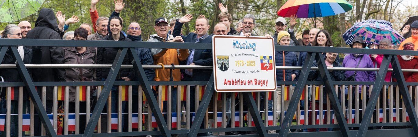 Viele Leute stehen mit dem Bürgermeister Florian A. Mayer auf einer Brücke mit dem Schild für die Partnerschaft mit Amberieu